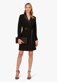 Robe noire cintrée à manches longues, col en satin et ceinture à nouer, associée à une pochette noire texturée et des talons noirs à lanières.