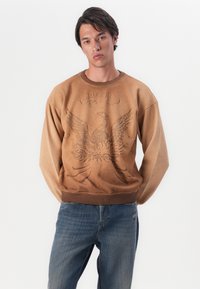 Bruine sweatshirt met reliëfafbeelding van een arend, geribbelde ronde hals en boorden. Gepaard met blauwe jeans. De stof vertoont een lichte textuur.