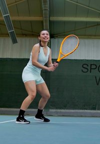 Joven mujer con un atuendo azul claro se prepara para golpear una pelota con una raqueta de tenis naranja en una cancha cubierta.