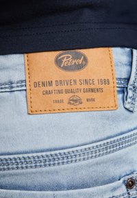 Blugi din denim albastru deschis, cu o etichetă din piele maro deschis inscripționată cu text negru: „Petrol DENIM DRIVEN DIN 1989 CREĂM HAINE DE CALITATE.”