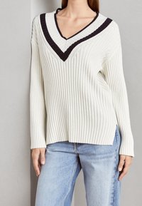 Kvinna klädd i en ribbad vit tröja med svarta halsremsor och ljusblå jeans, stående mot en enkel grå vägg.