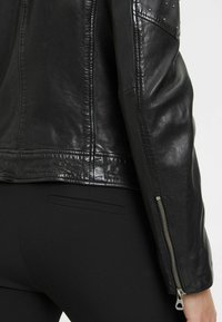 Giacca in pelle nera con finitura lucida, cuciture visibili, polsini con zip e dettagli in borchie decorative sulle spalle.