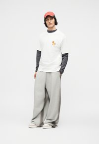 Νεαρό άτομο που φοράει ροζ καπέλο, λευκό μπλουζάκι με πορτοκαλί καρτούν στο στήθος, μακριά σκούρα μανίκια από μέσα, φαρδιά γκρι παντελόνια και λευκά αθλητικά παπούτσια.