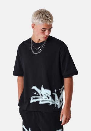 Joven con cabello rubio corto vistiendo camiseta negra de gran tamaño y pantalones cortos con diseños de graffiti blancos, mirando hacia abajo a su izquierda.