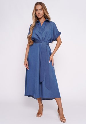 Kobieta z długimi włosami, ubrana w niebieską midi sukienkę z kołnierzykiem, krótkimi rękawami i paskiem wiązanym w talii, beżowe sandały na wysokim obcasie, stojąca.