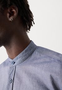 Galléros kék chambray ing finom textúrával, gombos nyakkal és kék gombokkal. Látható varrásokkal és klasszikus fazonban.