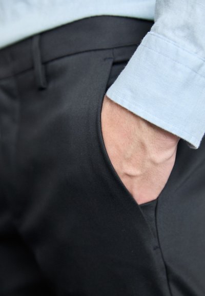Crne hlače s bočnim džepom, u kombinaciji s svijetloplavom košuljom s dugmadi na manžetama, s naglaskom na teksturu tkanine i prilagođenom dizajnu.