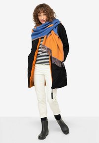 Zwarte, gevoerde jas met oranje voering, gestreepte top, crèmekleurige broek en zwarte enkellaarsjes. Blauw en oranje sjaal met franjes.