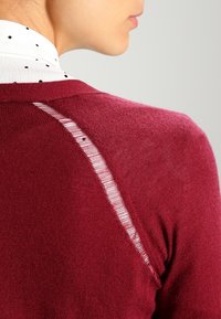 Burgundské pletené svetřík s kulatým výstřihem a jemným vertikálním pruhovaným detailem na zádech, který zdůrazňuje měkkou texturu a těsný střih.