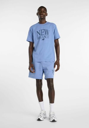 Homme portant un t-shirt New Balance bleu clair et un short, avec des chaussettes blanches et des chaussures de course, debout devant un fond blanc uni.