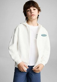 Felpa bianca con zip realizzata in fleece di cotone, con tasca frontale e logo blu. Indossata sopra una maglietta bianca e abbinata a pantaloni corti blu.