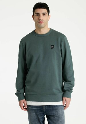 Joven con sudadera verde de cuello redondo con un pequeño logo negro en el pecho, superpuesta a una camisa blanca y vaqueros azules.