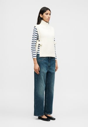 Κρεμ πλεκτό γιλέκο με ψηλό γιακά πάνω από μπλε σκούρο και λευκό ριγέ μακρυμάνικο τοπ, συνδυασμένο με φαρδιά μπλε τζιν παντελόνια και μαύρες μπαλαρίνες.
