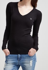 Černý dlouhý rukáv V-krk svetr vyrobený z měkkého a elastického materiálu. Na levé hrudi má malé trojúhelníkové logo. V kombinaci s ošuntělými modrými džíny.
