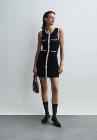 Kvinna med tatueringar klädd i en ärmlös svart klänning med vit kant, som håller en liten vinröd handväska, svarta ankelsockor och svarta klackskor.