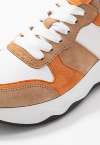 Baskets mettant en avant un mélange de cuir blanc, de daim orange et d'accents beige. Épaisses lacets blancs et semelle en caoutchouc texturée avec des détails noirs.