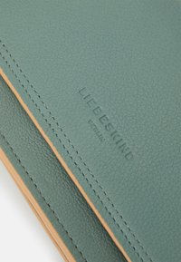 Portafoglio in pelle verde con texture, cuciture visibili e logo del marchio impressa. Presenta un dettaglio di bordo marrone chiaro lungo l'apertura.