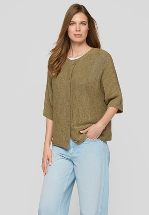 Frau mit langen Haaren, die eine olivgrüne, geknöpfte Strickjacke und hellblaue Jeans trägt, steht vor einem schlichten grauen Hintergrund.