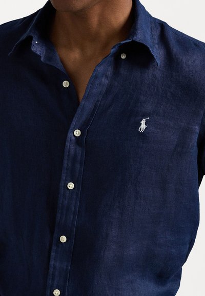 Jūras kara flotes linna pogu krekls ar klasisku apkakli, kuram ir baltas pogas un neliela izšūta logo kreisajā krūtī.