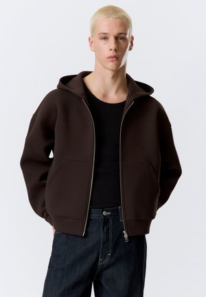 Jeune personne aux cheveux blonds courts portant un sweat à capuche zippé marron foncé sur un t-shirt noir et un jean foncé, se tenant devant un fond uni.