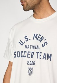 T-shirt branca de algodão com letras azul-escuro a dizer "U.S. Men's National Soccer Team 2026" e um pequeno emblema dos EUA na parte inferior.