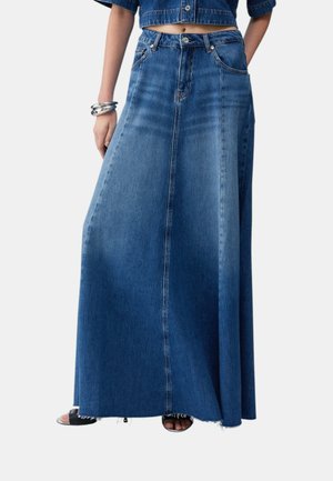 Gonna lunga di jeans blu con bottoni e cerniera frontali, indossata con un top di jeans corto, braccialetti argentati e sandali neri con punta aperta.