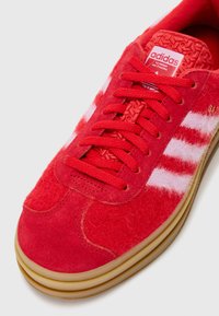 Sapatilha vermelha Adidas com riscas brancas felpudas, atacadores vermelhos, língua texturada e sola de borracha sobre fundo branco.