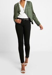 Mujer vestida con chaqueta verde de frente abierta, blusa blanca con botones, pantalones negros con cinturón y tacones blancos con tira en el tobillo caminando hacia adelante.
