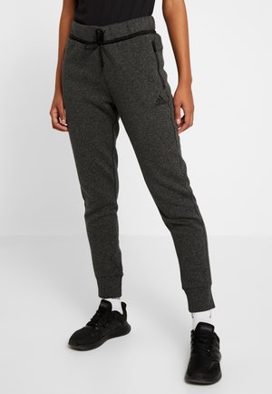Personne portant un pantalon de survêtement Adidas gris foncé avec une taille à cordon et des baskets noires, debout sur un fond blanc uni.