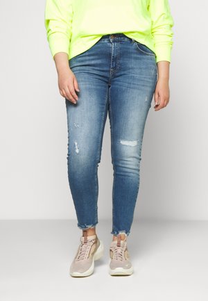 Personne portant un jean skinny bleu clair usé avec des ourlets effilochés, un haut à manches longues jaune néon et des baskets rose clair, debout sur un sol gris.