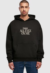 Schwarzer Kapuzenpullover aus weichem Stoff mit einer Kängurutasche. Der Grafikdruck auf der Vorderseite zeigt "tupac ALLEYEZ ON ME" in Beige.