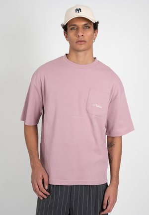 Jonge man draagt een lichtroze kortemouwen shirt met borstzak en donkere fijn gestreepte broek, beige pet met logo, staand voor een witte achtergrond.