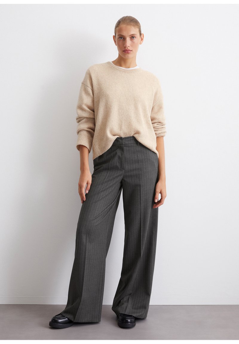 Beige strikkegenser med en løs passform og ribbede mansjetter, kombinert med mørkegrå bukser med pinstriper og svarte sko.