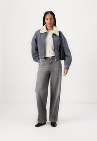 Grå kordfløyelsjakke med en kremfarget shearlingkrage, beige genser og løse grå jeans. Modell holder en svart håndveske og har på seg svarte sko.