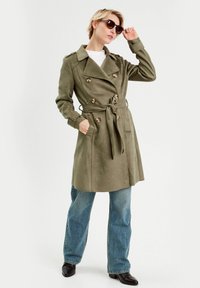 Trench coat verde oliva con botones de doble fila, cinturón en la cintura y hombreras, combinado con jeans acampanados azules y zapatos negros.