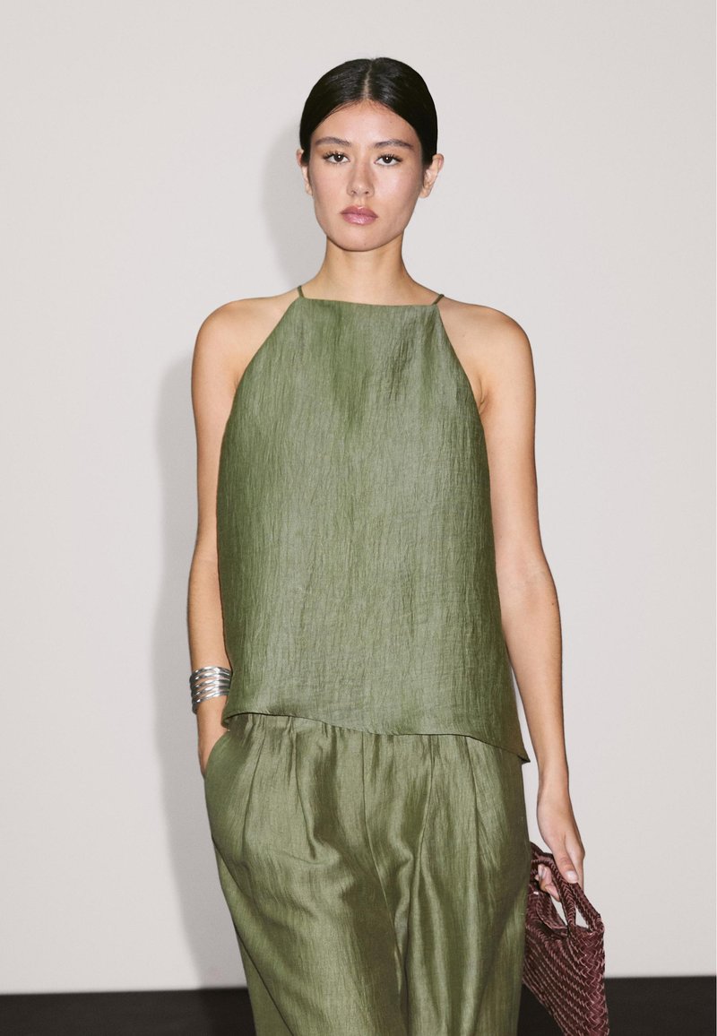 Green linen halter top with a relaxed fit, featuring textured fabric and a slightly curved hem, paired with matching pants and a burgundy bag.