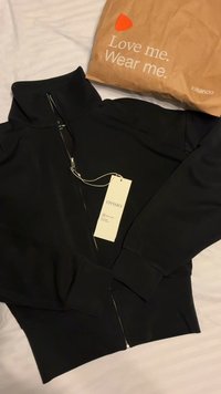 Černá mikina na zip s tagem Oysho ležící na bílé podložce vedle hnědého papírového sáčku Zalando s textem "Miluj mě. Nosi mě."