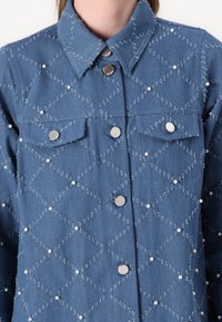 Denimjakke med diamantmønster, sølvperler, trykknapper og to brystlommer; fargen er medium blå med teksturert overflate.