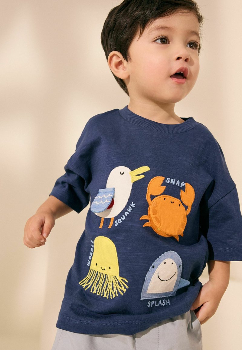 Ragazzo giovane che indossa una camicia blu navy con toppe colorate di animali marini e parole onomatopeiche, guarda di lato con la bocca leggermente aperta.