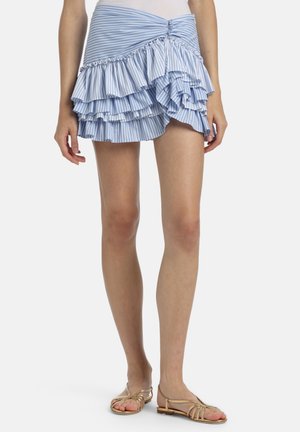 Mujer con una falda mini de volantes a rayas azules y blancas y sandalias planas doradas con tiras, de pie contra un fondo liso.