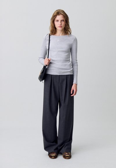 Maglia grigio a maniche lunghe con colletto rotondo, abbinata a pantaloni larghi a righe. Il modello tiene una piccola borsa nera; accessori non evidenti.