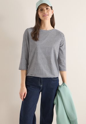 Femme portant une casquette bleu clair, un haut à manches longues rayé gris et blanc, un jean foncé, tenant un pull vert menthe sur un fond beige.