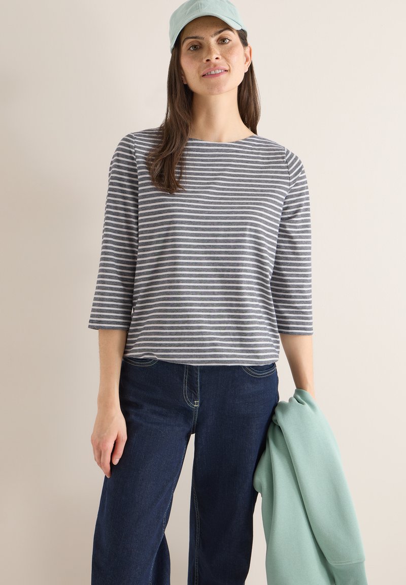 Femme portant une casquette bleu clair, un haut à manches longues rayé gris et blanc, un jean foncé, tenant un pull vert menthe sur un fond beige.