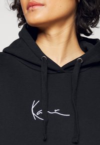 Czarna bluza z kapturem uszyta z miękkiego materiału, z białym haftowanym logo na klatce piersiowej, z regulowanymi sznurkami i metalowymi oczkami.