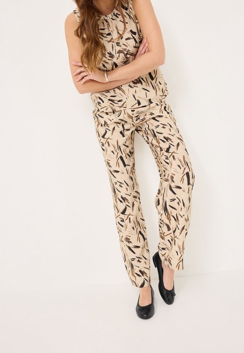 Femme portant un haut beige sans manches et un pantalon assorti avec des motifs abstraits de coups de pinceau noirs et bruns, debout contre un mur blanc.