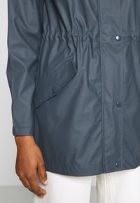 Imperméable bleu foncé avec fermeture à boutons-pression sur le devant et poche à rabat inclinée, porté sur un pantalon blanc, montrant le bras et la main gauche.