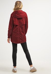 Czerwona kurtka z kapturem o luźnym kroju, z tylną częścią w stylu peleryny, z lekkiego materiału i głębokimi bocznymi kieszeniami, zestawiona z dopasowanymi czarnymi spodniami.