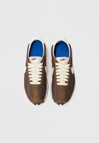 Brūni sporta apavi ar tīkla un zamšādas materiāliem, baltiem šņorēm, ziliem zolēm un izceļošu Nike logo uz mēles. Noapaļota purngala dizains.