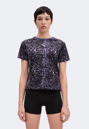 Ung person med kort mørkt hår iført en lilla og sort leopardprint t-shirt og sorte shorts, stående mod en ensfarvet baggrund.