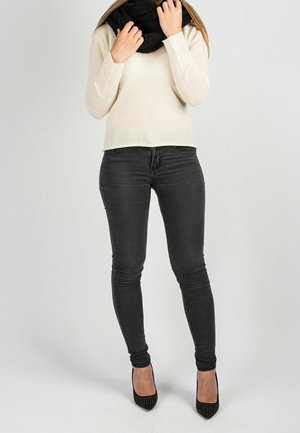 Femme portant un pull blanc, une écharpe noire, un jean skinny sombre et des talons hauts noirs cloutés, debout devant un fond gris uni.